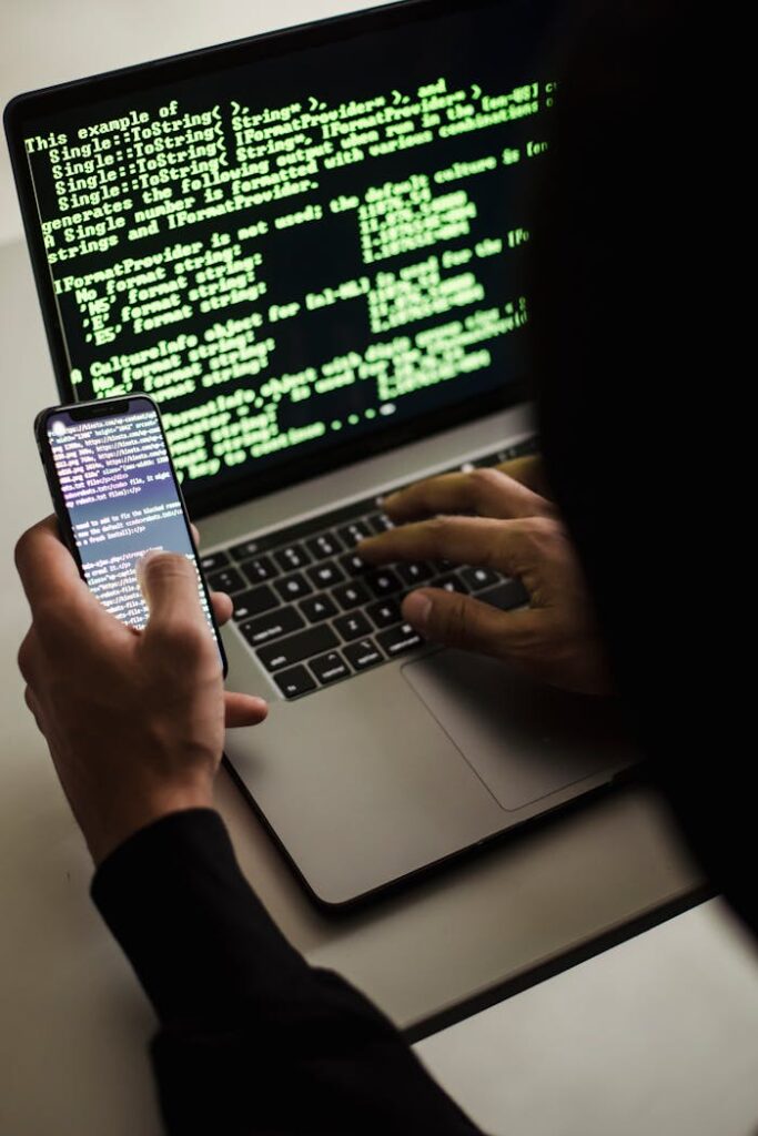 pexels-photo-5935791 A hooded figure engaged in hacking using a laptop and smartphone in low light.